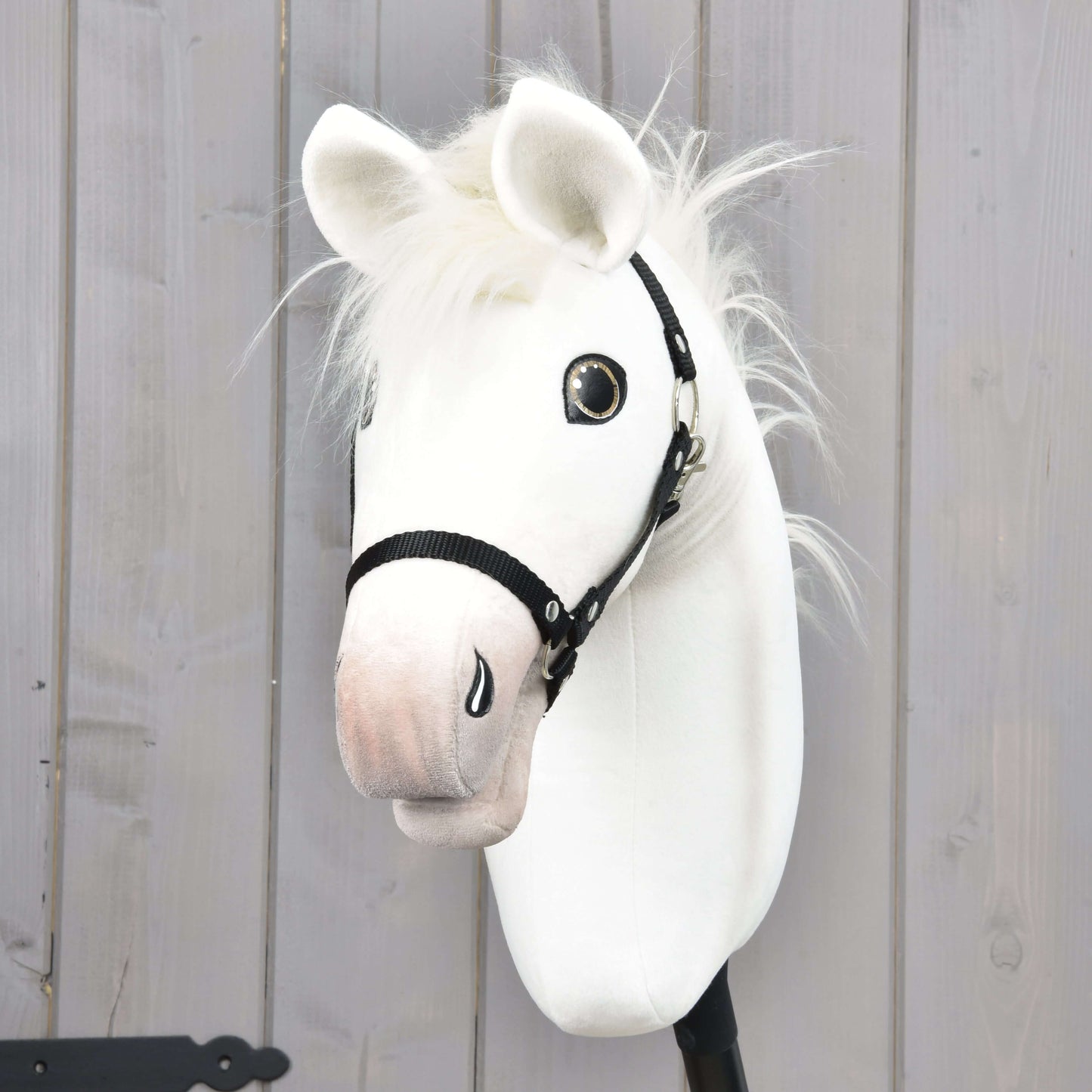 The Hobby Horse Nexus with Black Halter by Hobby Horse LarDen, a white fabric hobby horse with a soft mane and shiny eyes, is shown against a light wooden panel background.
