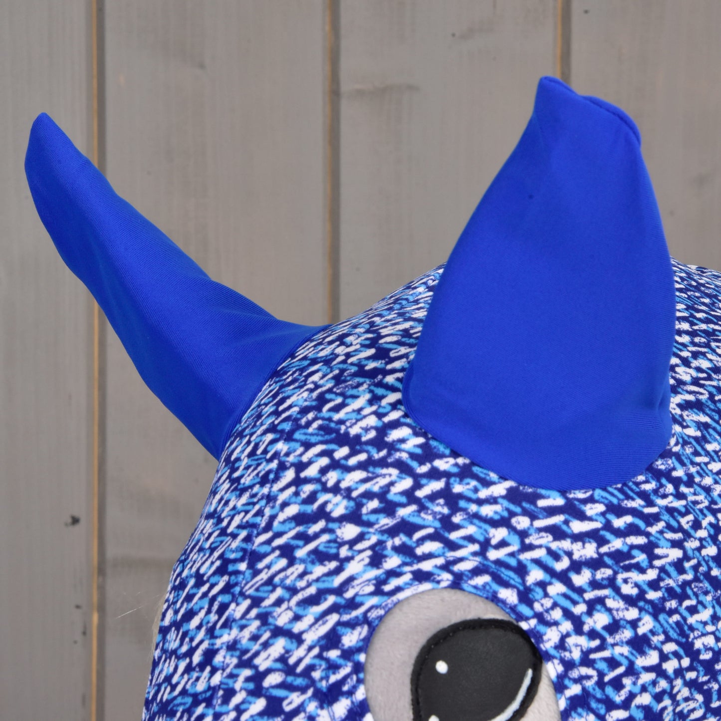 A close-up of the Hobby Horse LarDen Hobby Horse protective suit See, made from textured blue PES material with two large blue horns and a round cartoon eye, shown against a wooden background.