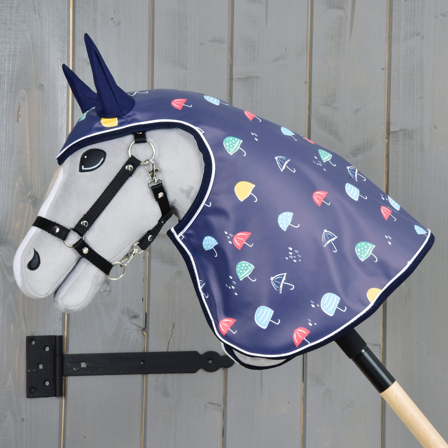 The Hobby Horse LarDen "Raincoat Rainy" features a plush horse with a bridle and blue unicorn horn, wearing a navy raincoat with colorful umbrellas, mounted on a wooden stick against a gray wood background—perfect for all-weather play.