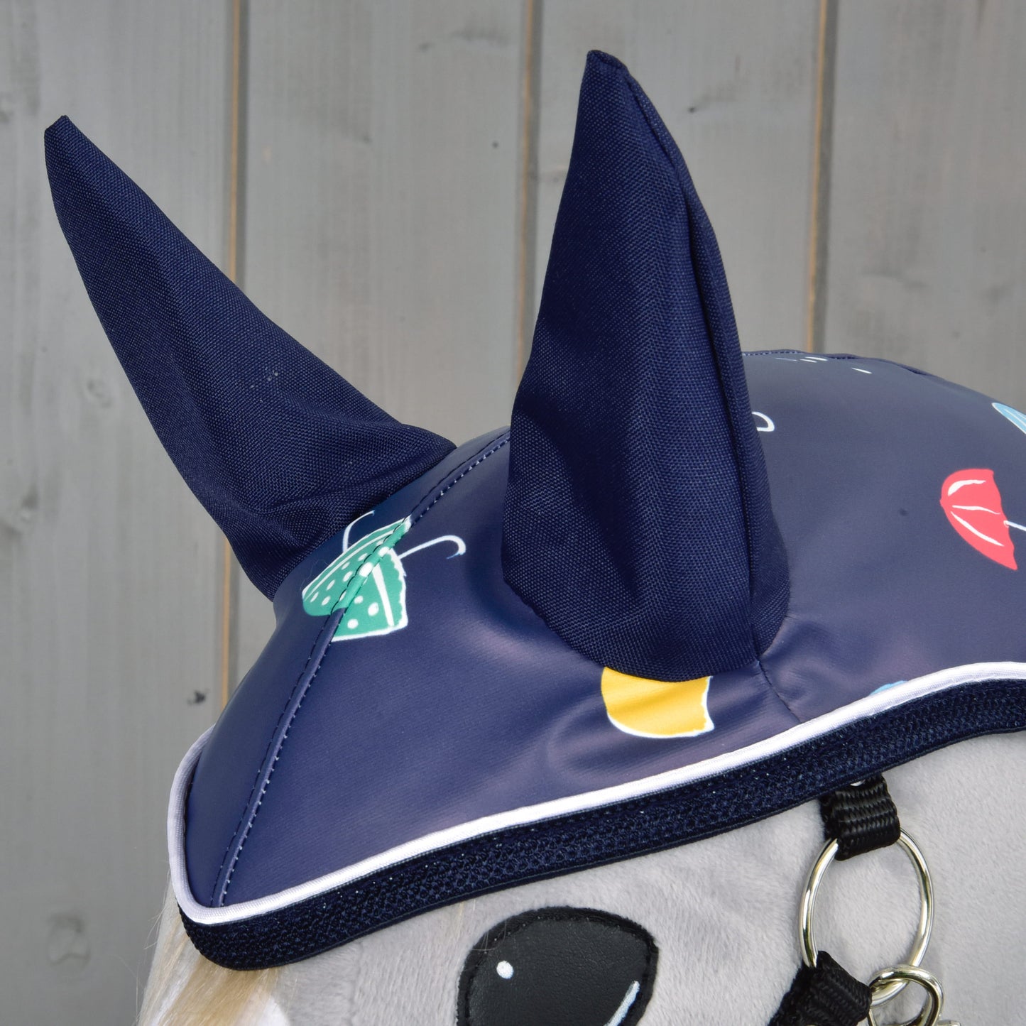 Close-up of a Hobby Horse LarDen’s Raincoat Rainy bonnet in blue, featuring upright ears and vibrant umbrella and raindrop prints—ideal for any weather. Set against a light gray wooden wall backdrop.