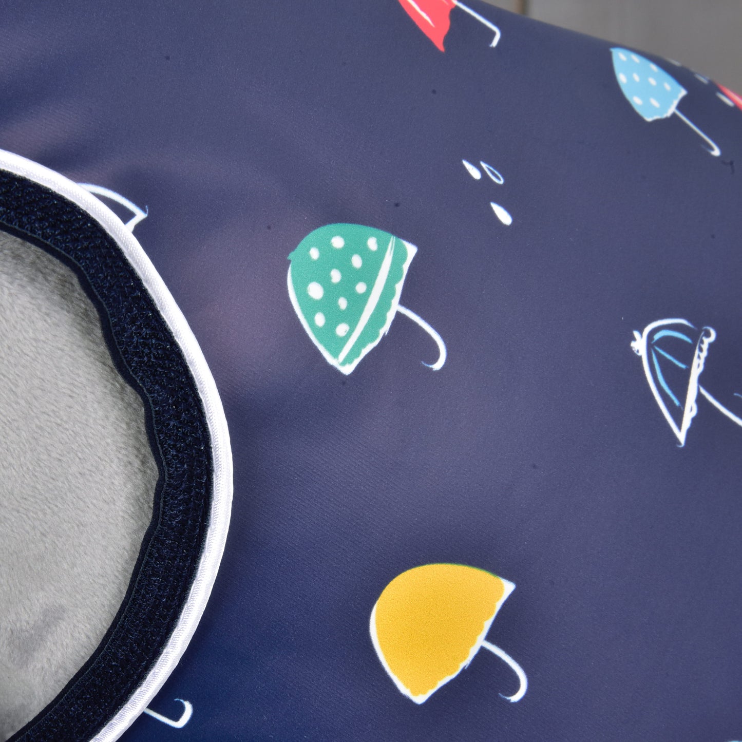 A close-up of Raincoat Rainy by Hobby Horse LarDen shows navy blue fabric with green, yellow, red, and blue umbrellas and a soft grey lining visible on the left side—perfect for all weather.