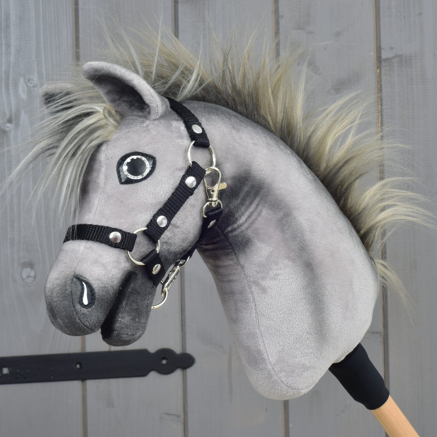 The Hobby Horse LarDen “Hobby Horse Prestige with black halter, size XS” features a gray plush design with a spiky mane and black halter, inspired by the elegance of an English Warmblood. It is mounted on a wooden stick and displayed against a wooden panel background.