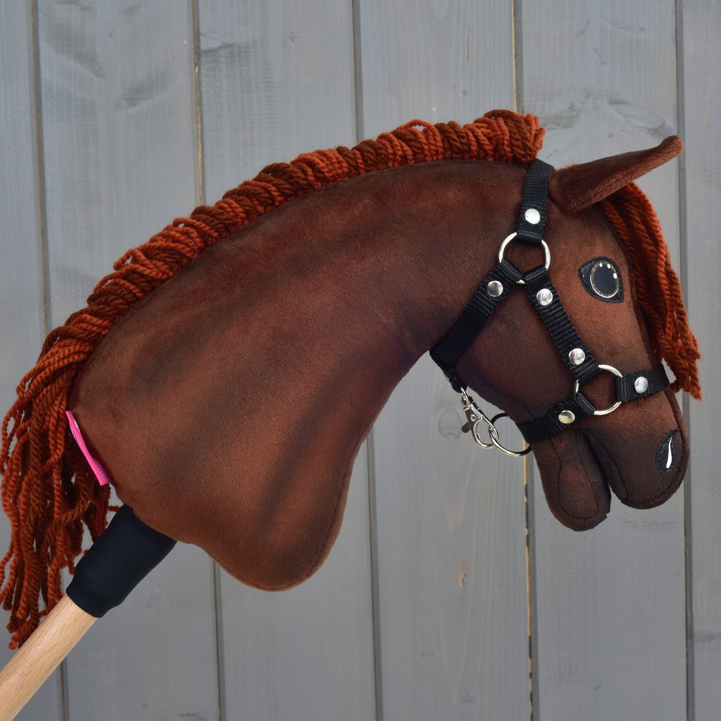 A Hobby Horse LarDen "Rebel" hobby horse in size XS, featuring a brown Hanoverian design with a yarn mane and black halter, is positioned in front of a gray wooden background.