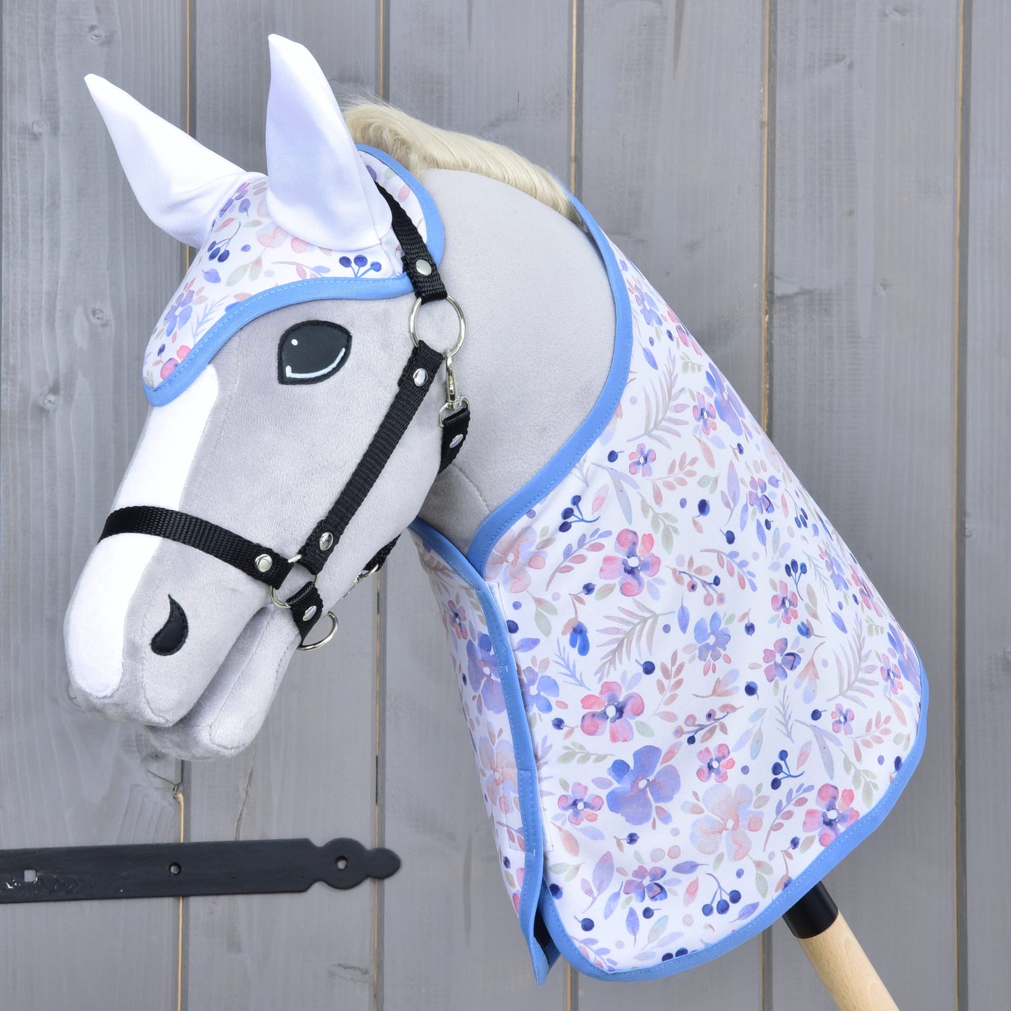 Set Vanessa NEW de Hobby Horse LarDen, avec Hobby Horse gris, bride noire, ensemble couverture floral à bordure bleue et bonnet d'oreilles assorti, présenté sur fond en bois.