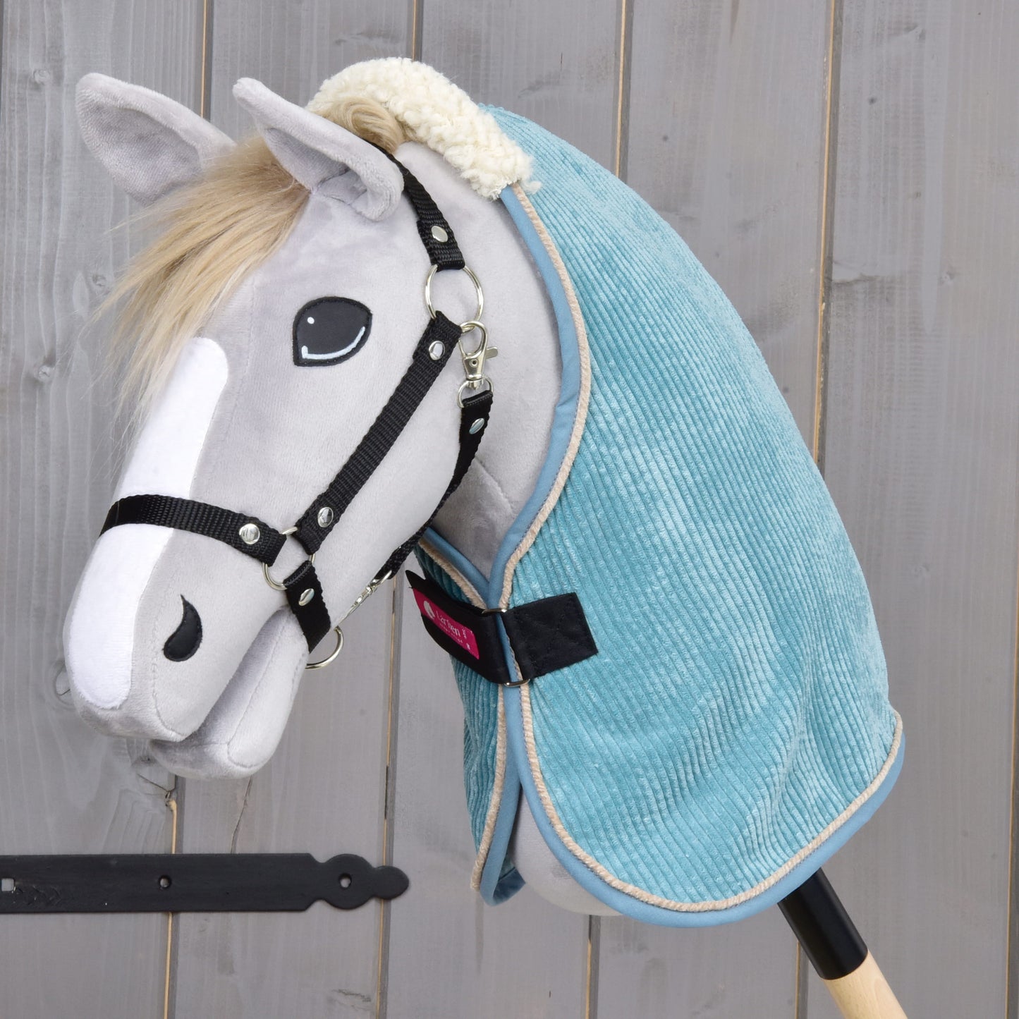A plush hobby horse by Hobby Horse LarDen features a light gray face, black bridle, white muzzle, and wears the Stable Blanket Fern in light blue PES material, posed against a wooden background.