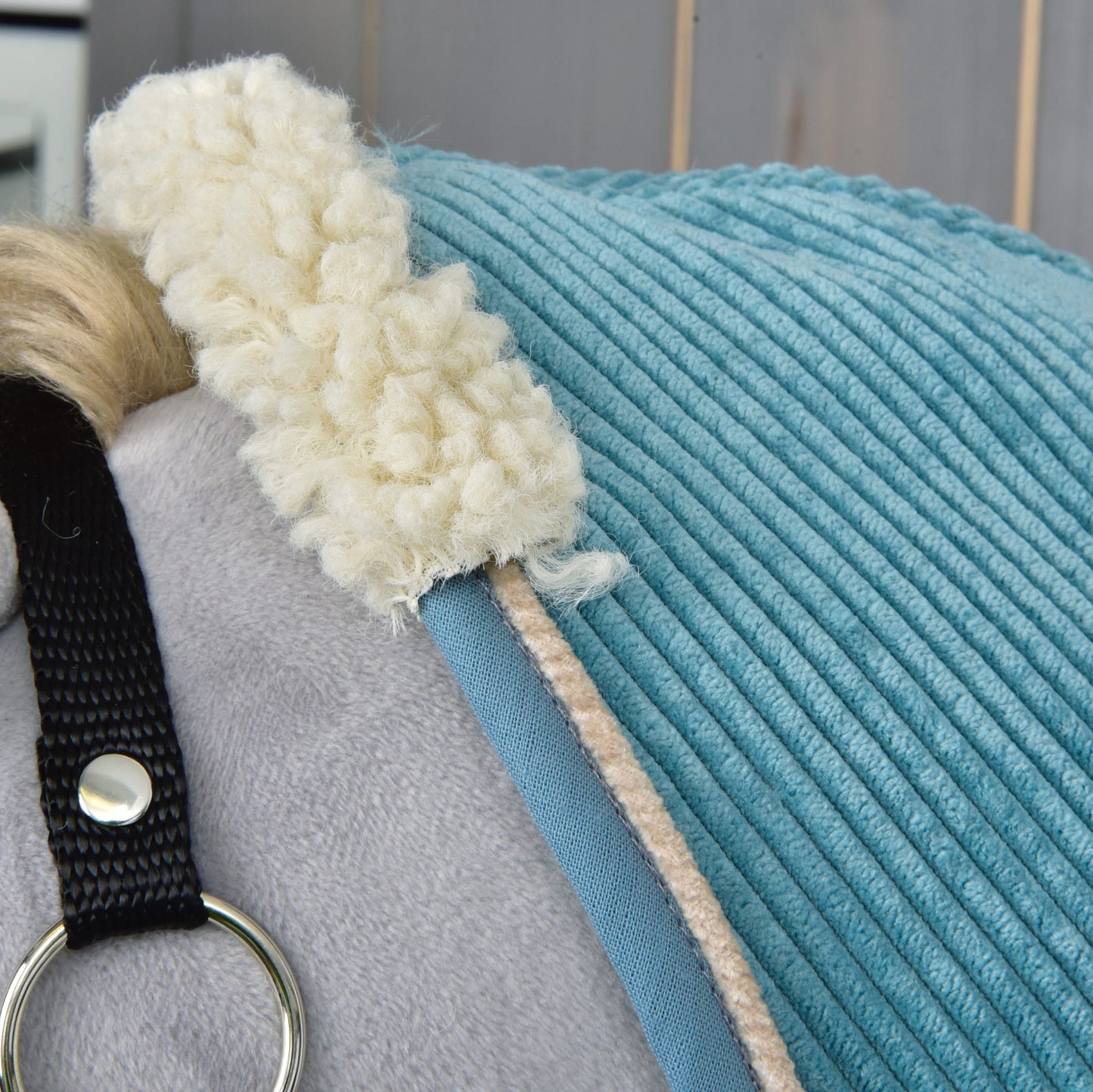 Close-up of a light grey plush horse wearing the blue Stable Blanket Fern by Hobby Horse LarDen, featuring cream fleece trim and a black strap with silver ring. Light wooden panels are visible in the background.