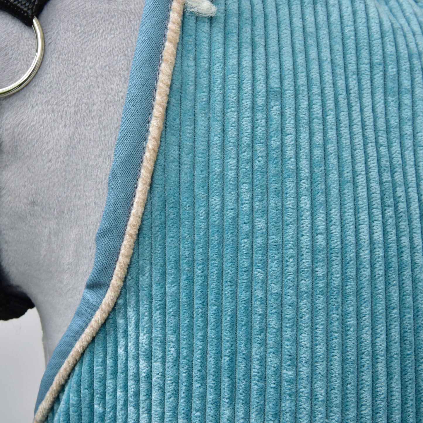 Close-up of a textured blue Stable Blanket Fern by Hobby Horse LarDen draped over a gray horse, featuring a beige fuzzy edge and a visible metal ring from the horse’s harness.