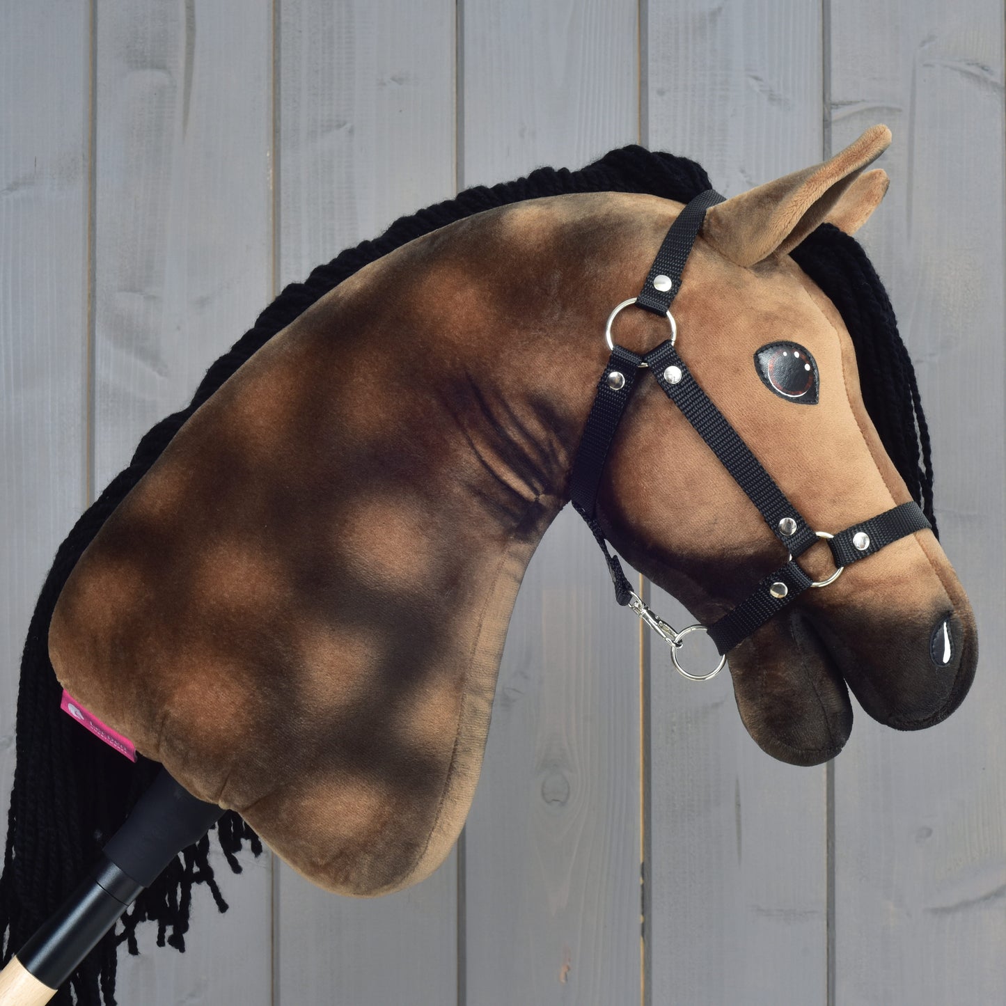 The Hobby Horse LarDen Hobby Horse Stormy with Black Halter—a plush dapple grey hobby horse with a brown and black mane—is displayed in front of a wooden panel background.