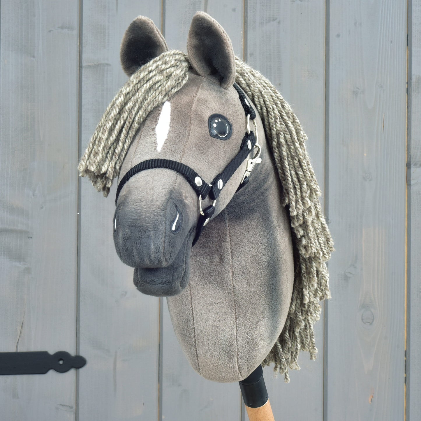 A close-up of the Hobby Horse LarDen Hobby Horse Swing with black halter, size S, shows its elegant gray design with a yarn mane, button eyes, a distinctive white blaze down the face, and a black halter—evoking the refined look of a Hanoverian stallion against a wooden background.