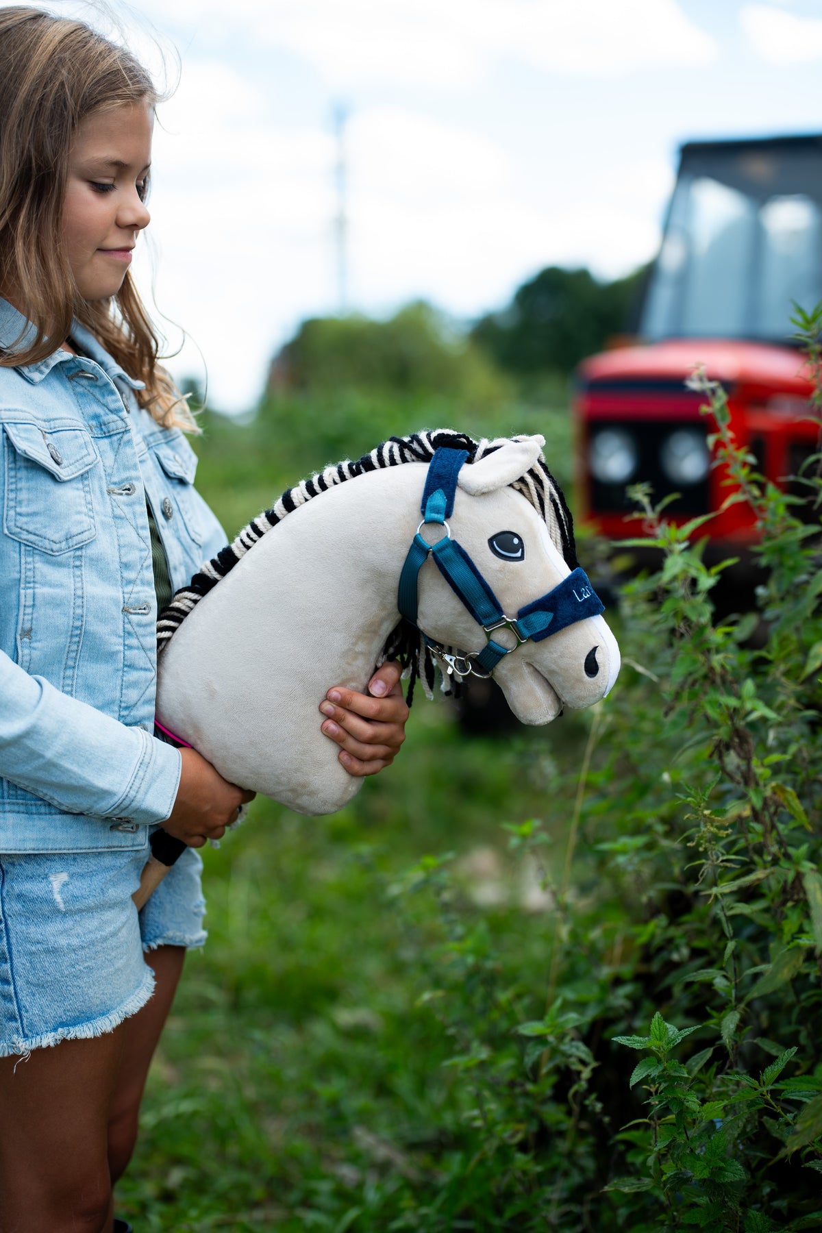 Une fille en veste en jean tient le Hobby Horse Aurora avec Licol noir de Hobby Horse LarDen près d’une haie, un tracteur rouge flou à l’arrière-plan, tête en peluche blanc crème et crinière en fil noir et blanc rayé, avec le licol LarDen Blue doublé de fourrure
