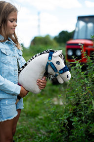 Une fille en veste en jean tient le Hobby Horse Aurora avec Licol noir de Hobby Horse LarDen près d’une haie, un tracteur rouge flou à l’arrière-plan, tête en peluche blanc crème et crinière en fil noir et blanc rayé, avec le licol LarDen Blue doublé de fourrure
