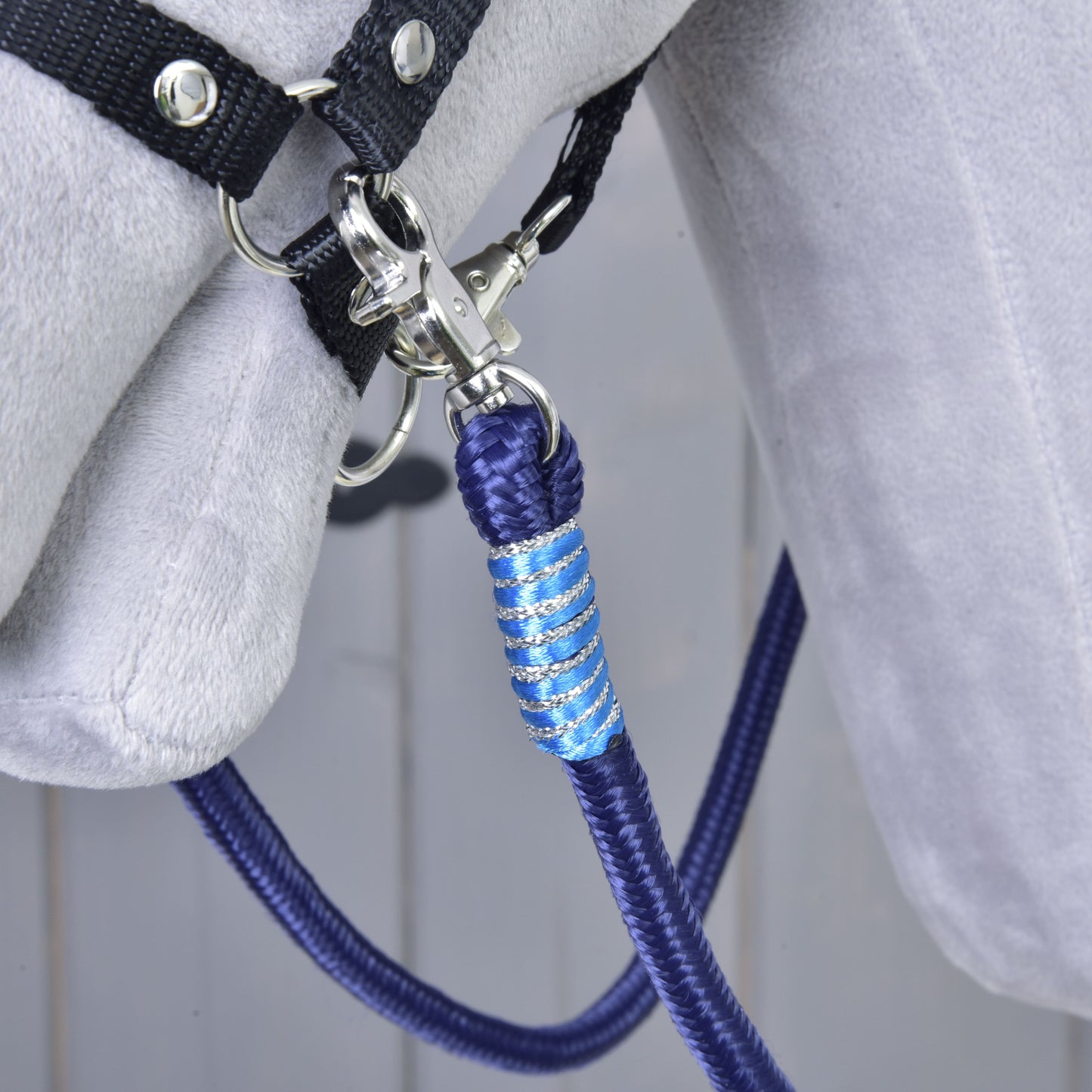 A close-up of the Hobby Horse LarDen Reins Blues, a blue and silver braided polypropylene lead rope, attached to a black halter on a grey horse against a wooden background.