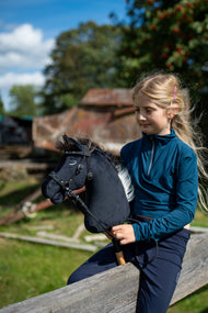 Une fille blonde en haut bleu sarcelle est assise sur une barrière en bois dans une cour de ferme ensoleillée, tenant le Hobby Horse Bond avec Licol noir de Hobby Horse LarDen, tête en peluche noire et crinière en fil marron foncé, avec la bride sans mors décorée
