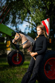 Une fille en col roulé noir se tient près d’une machine agricole verte en tenant le Hobby Horse Bren avec Licol noir de Hobby Horse LarDen, tête en peluche marron sable et crinière en fil crème, avec la bricole Espresso avec fourrure
