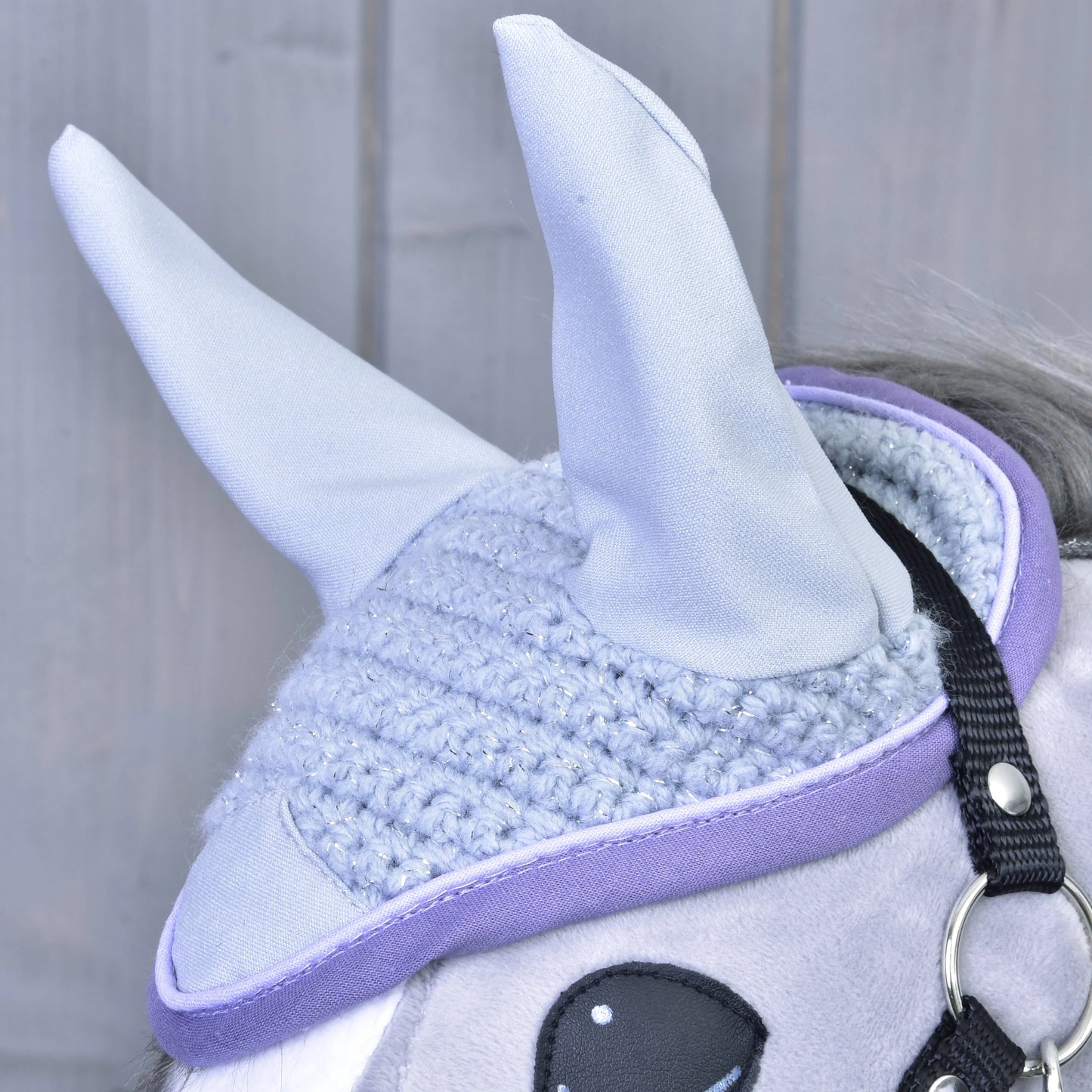 Close-up of Hobby Horse LarDen’s Ear bonnet crocheted Candy on a plush horse toy. The light purple bonnet with upright fabric ears provides insect protection. Gray wooden panels form the background.