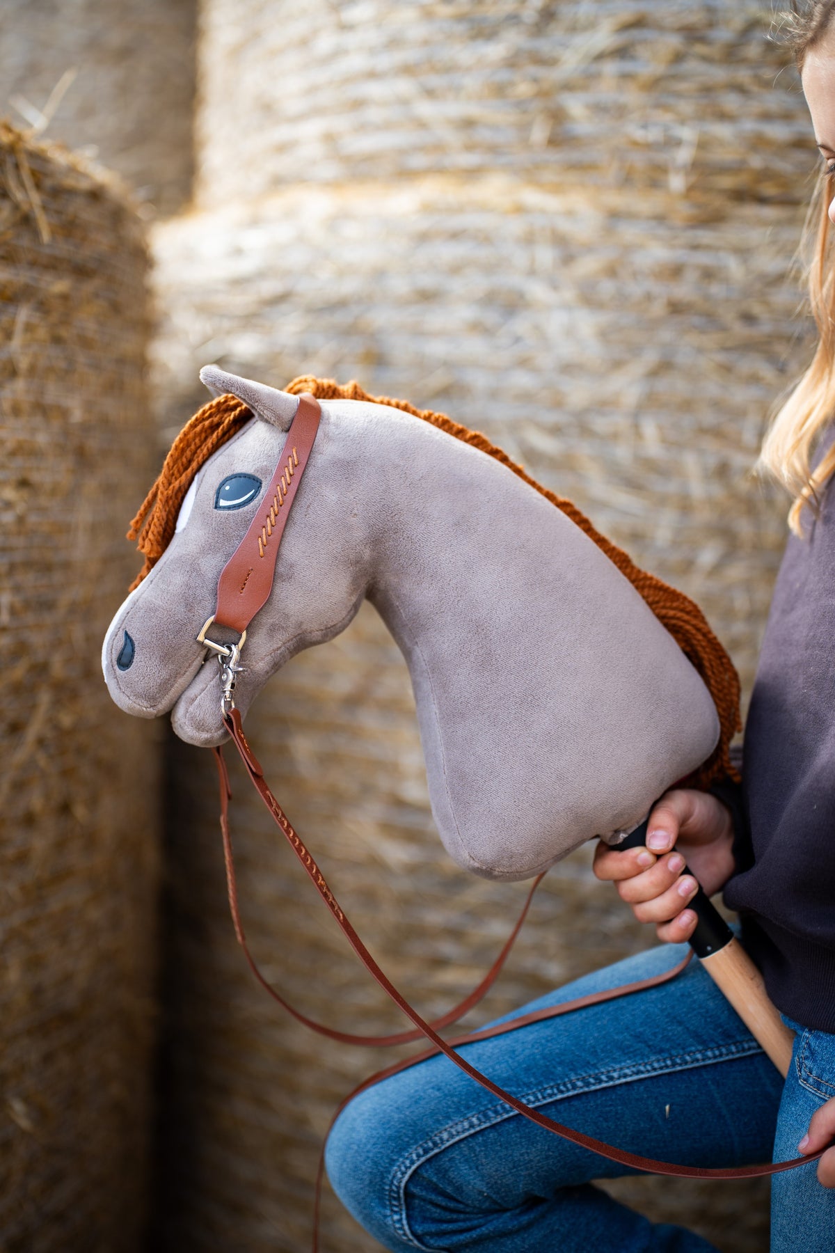 La tête du Hobby Horse Chanell avec Licol noir de Hobby Horse LarDen devant des balles de paille rondes, peluche grise, œil bleu et crinière en fil roux, avec la bride Western complète à une oreille en cuir marron