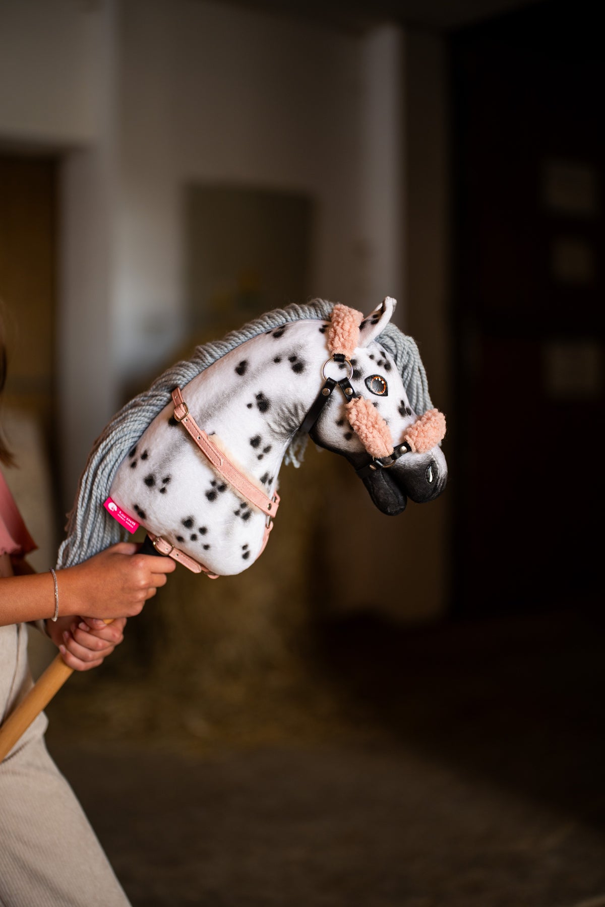 La tête et l’encolure du Hobby Horse Chantal avec Licol noir de Hobby Horse LarDen tenue dans une écurie, peluche blanche à taches léopard noires, crinière en fil gris et museau noir, avec le licol Old Rose en similicuir avec fourrure et la bricole avec fourrure