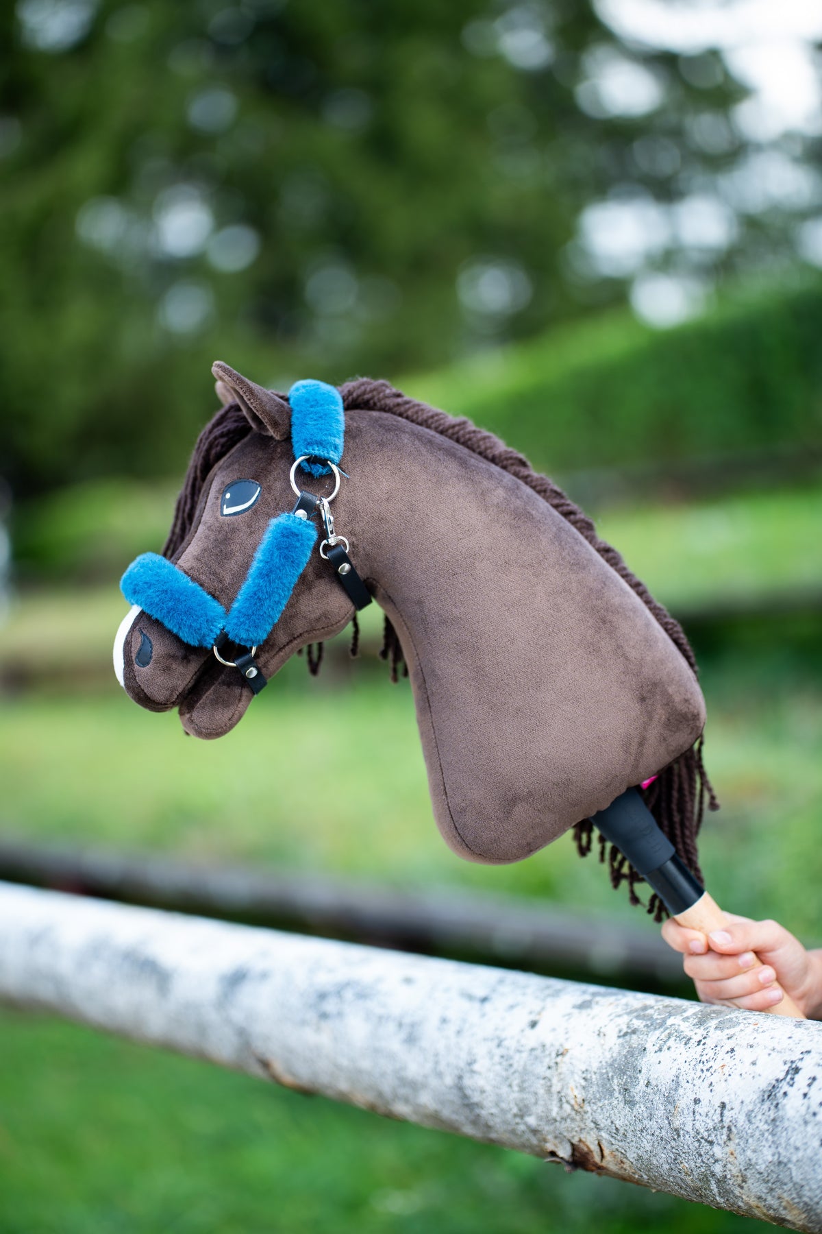 La tête du Hobby Horse Chianti avec Licol noir de Hobby Horse LarDen posée sur une barrière blanche de paddock, peluche marron foncé, crinière et toupet en fil foncé, avec le licol Teddy Petrol doublé de fourrure bleu vif