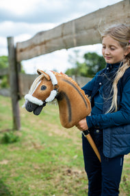 Une fille en gilet bleu marine se tient près d’une clôture de paddock en bois en tenant le Hobby Horse Connie avec Licol noir de Hobby Horse LarDen, tête en peluche alezane avec raie dorsale foncée et crinière en fil crème, avec le licol Teddy Classic doublé de fourrure