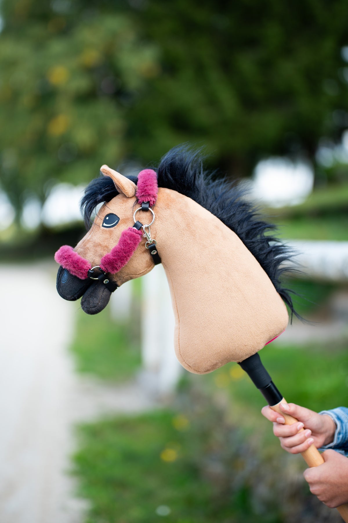 La tête du Hobby Horse Cookie avec Licol noir de Hobby Horse LarDen tenue près d’un chemin bordé d’arbres, peluche sable et longue crinière en fil noir, avec le licol Teddy Maroon doublé de fourrure