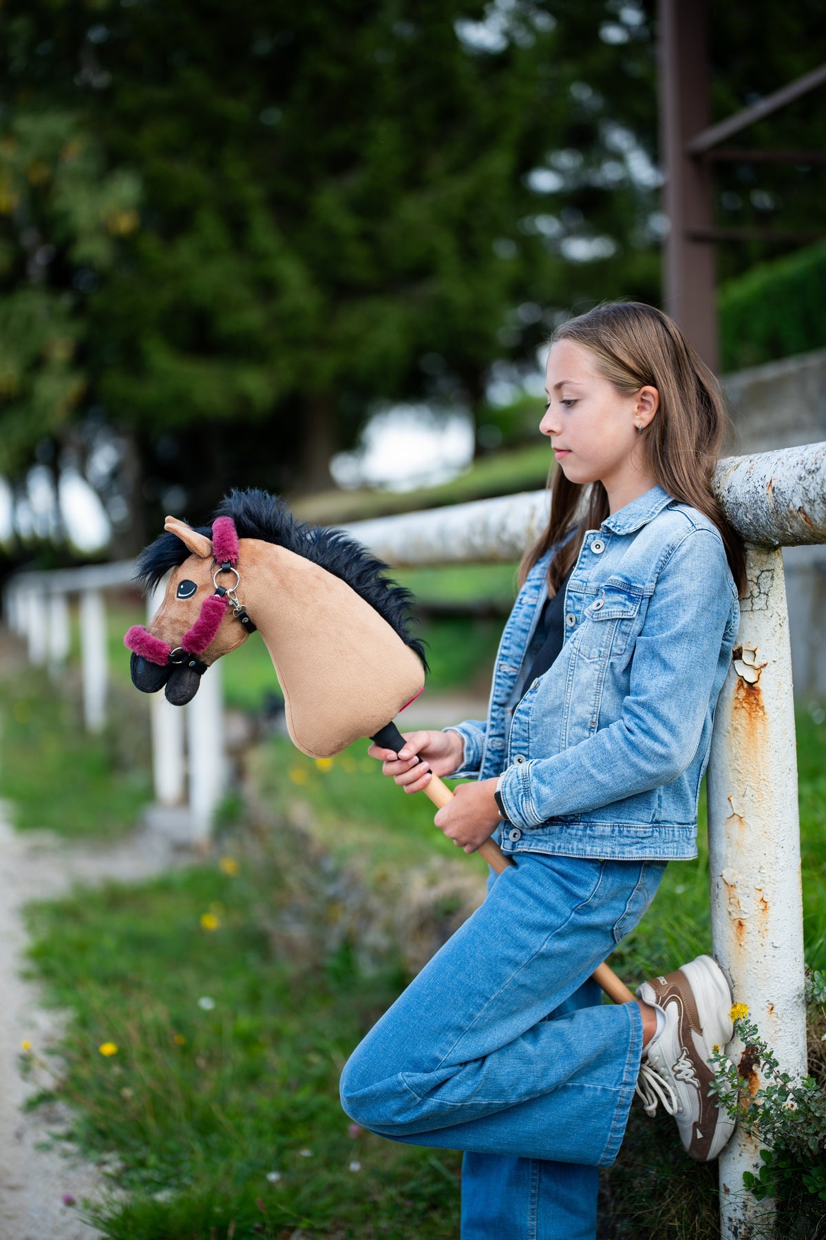 Une fille en tenue tout jean s’appuie contre une barrière blanche de paddock en tenant le Hobby Horse Cookie avec Licol noir de Hobby Horse LarDen, tête en peluche sable et crinière en fil noir, avec le licol Teddy Maroon doublé de fourrure