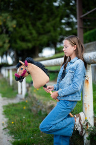 Une fille en tenue tout jean s’appuie contre une barrière blanche de paddock en tenant le Hobby Horse Cookie avec Licol noir de Hobby Horse LarDen, tête en peluche sable et crinière en fil noir, avec le licol Teddy Maroon doublé de fourrure