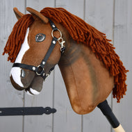 Hobby Horse Cooper avec licol noir de Hobby Horse LarDen, Hobby Horse brun inspiré d’un cheval de sport Oldenbourg. Ce jouet présente une liste blanche sur le visage, une épaisse crinière en fil brun foncé et est livré avec un licol en nylon noir orné de ferrures métalliques. Fixé à un bâton en bois et photographié devant des planches de bois grises, il est idéal pour le jeu imaginatif de dressage.