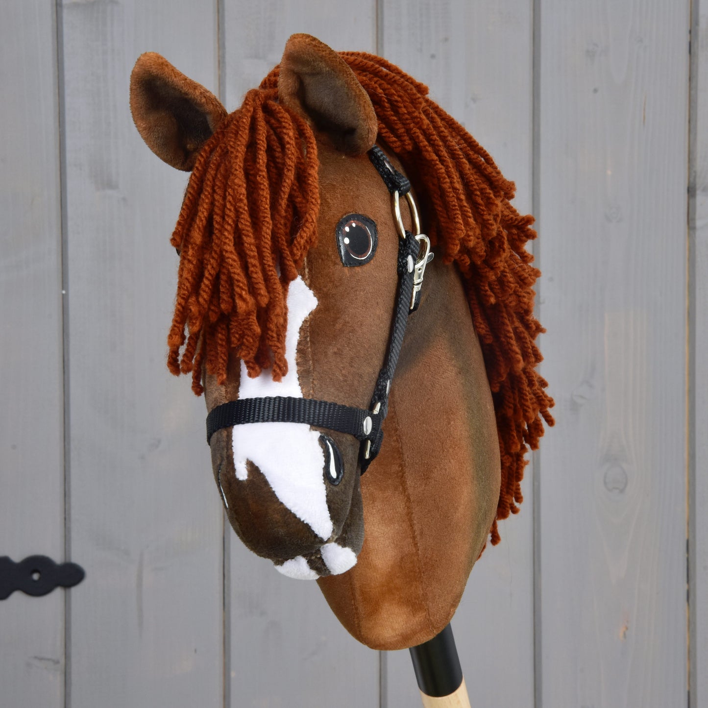The Hobby Horse LarDen "Hobby Horse Cooper with Black Halter" features a brown plush design, white facial markings, and a thick yarn mane inspired by the elegant Oldenburg sport horse, displayed against a wooden background.