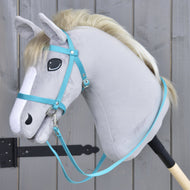 A gray plush hobby horse with a light mane, wearing the Hobby Horse LarDen black bridle with reins from strap turquoise, is mounted on a wooden stick and placed before a gray wooden fence.
