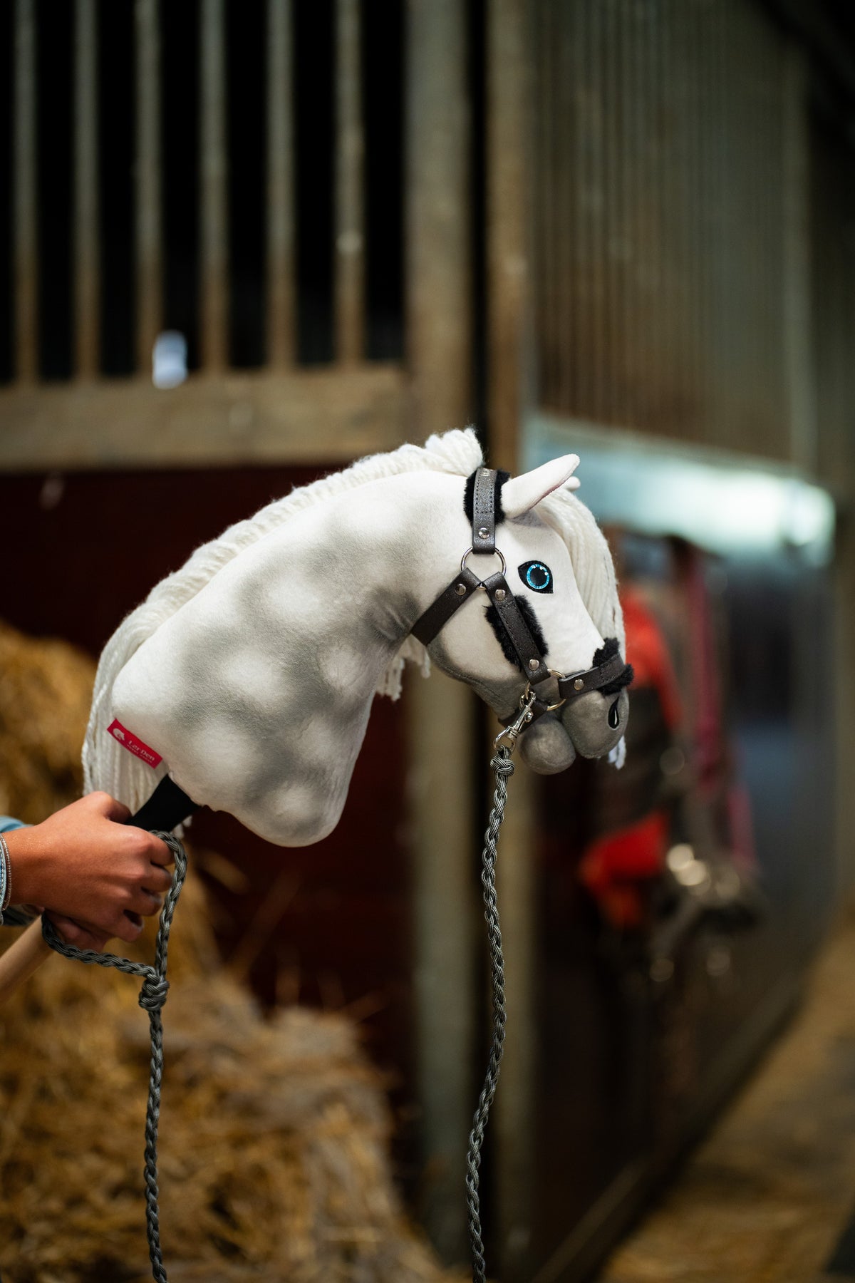 La tête du Hobby Horse Elektra avec Licol noir de Hobby Horse LarDen tenue près des boxes, un cheval couvert flou derrière, peluche blanche pommelée, œil bleu et crinière en fil blanc, avec le licol Graphite Full en similicuir avec fourrure et la longe tricotée