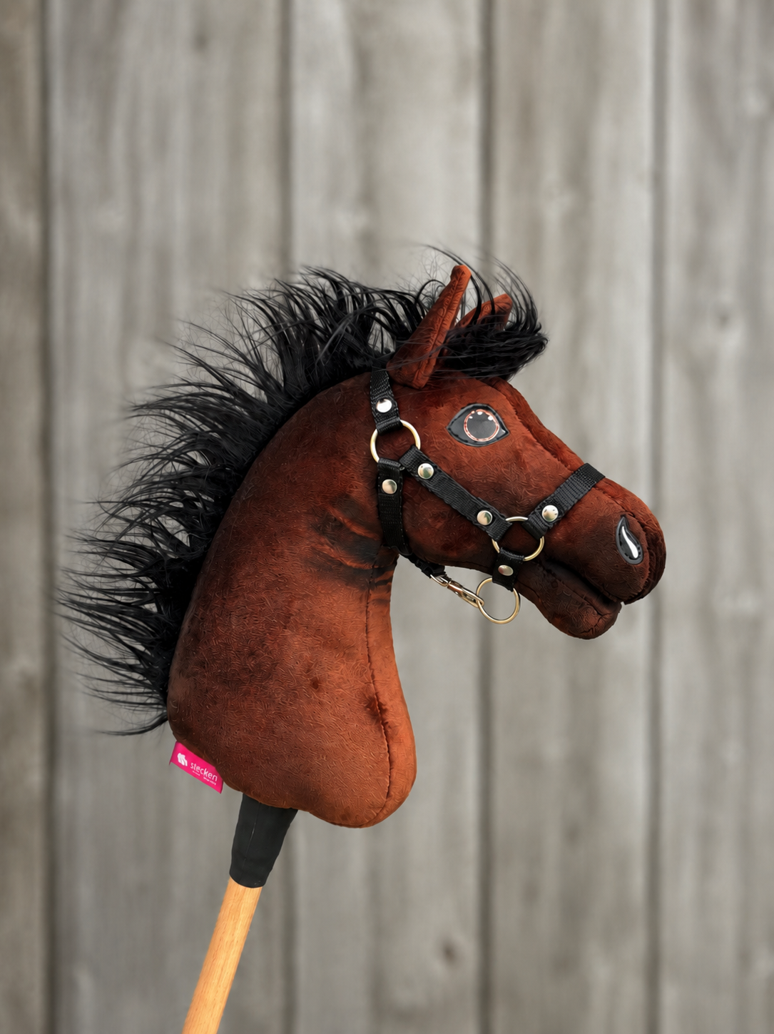 The Hobby Horse LarDen "Hobby Horse Benny with Black Halter" (size XS) features a brown plush body, black mane, and sits on a wooden stick. Shown against a blurred wooden plank background, it closely resembles a sport horse.