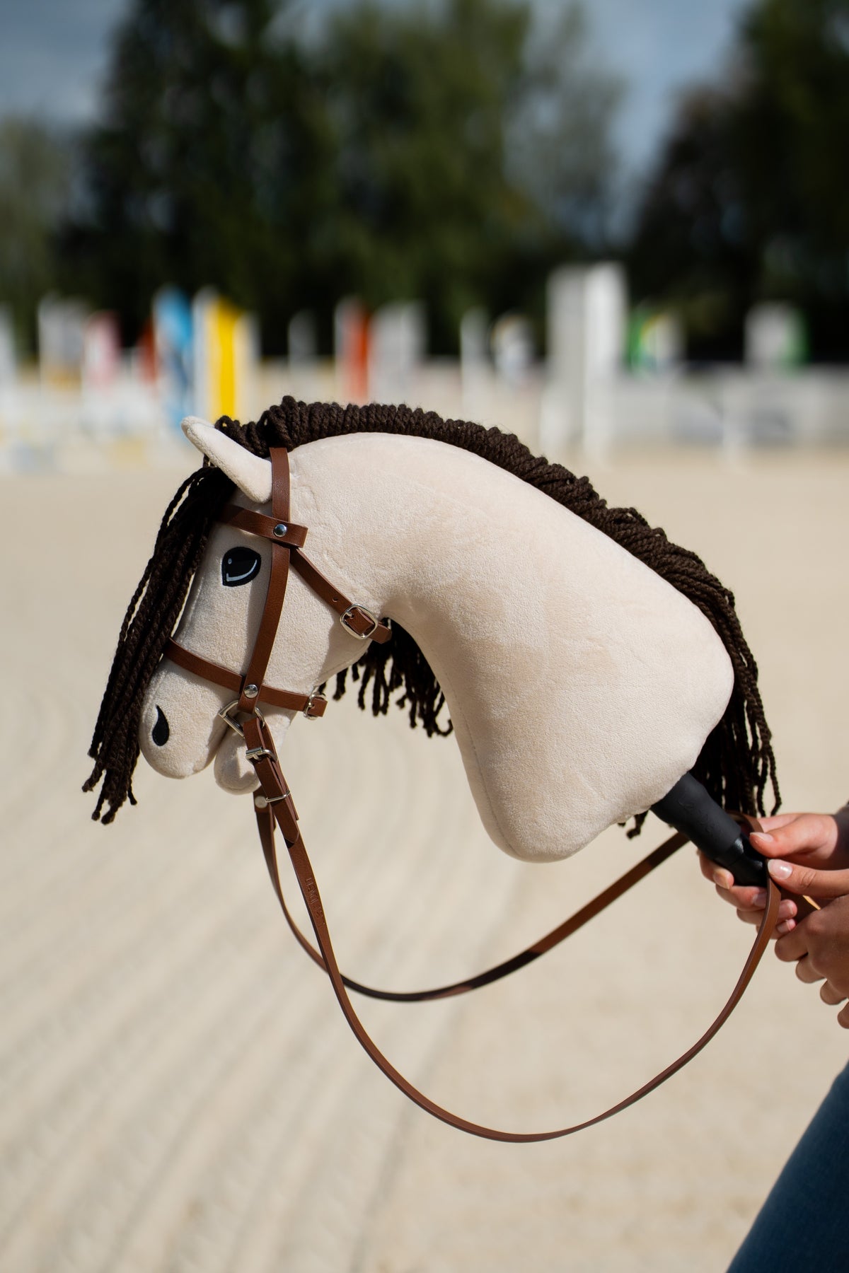 Hobby Horse Frederik LD crème à crinière en laine marron foncé avec bride marron, muserolle et longues rênes, obstacles en arrière-plan