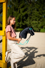 Une fille en haut rose s’appuie contre un chandelier d’obstacle jaune en tenant le Hobby Horse Freya avec Licol noir de Hobby Horse LarDen, tête en peluche noire, liste blanche et crinière en fil noir, avec la bride turquoise complète sans muserolle