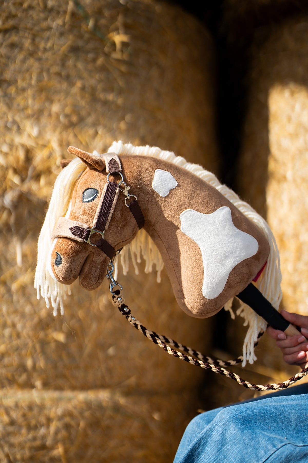 La tête et l’encolure du Hobby Horse Fury avec Licol noir de Hobby Horse LarDen devant des balles de paille rondes, peluche palomino à marques blanches et crinière en fil crème, avec le licol LarDen Brown doublé de fourrure et la longe tressée Bloom Brown