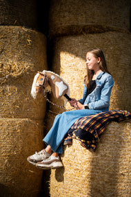 Une fille en veste en jean est assise sur de hautes balles de paille rondes en tenant le Hobby Horse Fury avec Licol noir de Hobby Horse LarDen, tête en peluche palomino à marques blanches et crinière en fil crème, avec le licol LarDen Brown doublé de fourrure et la longe Bloom Brown
