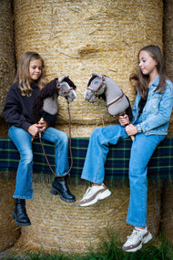 Deux filles sont assises sur des balles de paille recouvertes d’une couverture écossaise, chacune tenant un Hobby Horse Guccini de Hobby Horse LarDen, tête grise de style Holsteiner avec liste blanche sur le nez et longue crinière en fil marron foncé, présenté avec le licol Walnut doublé de fourrure, la bricole Walnut avec fourrure et la longe Vivaldi