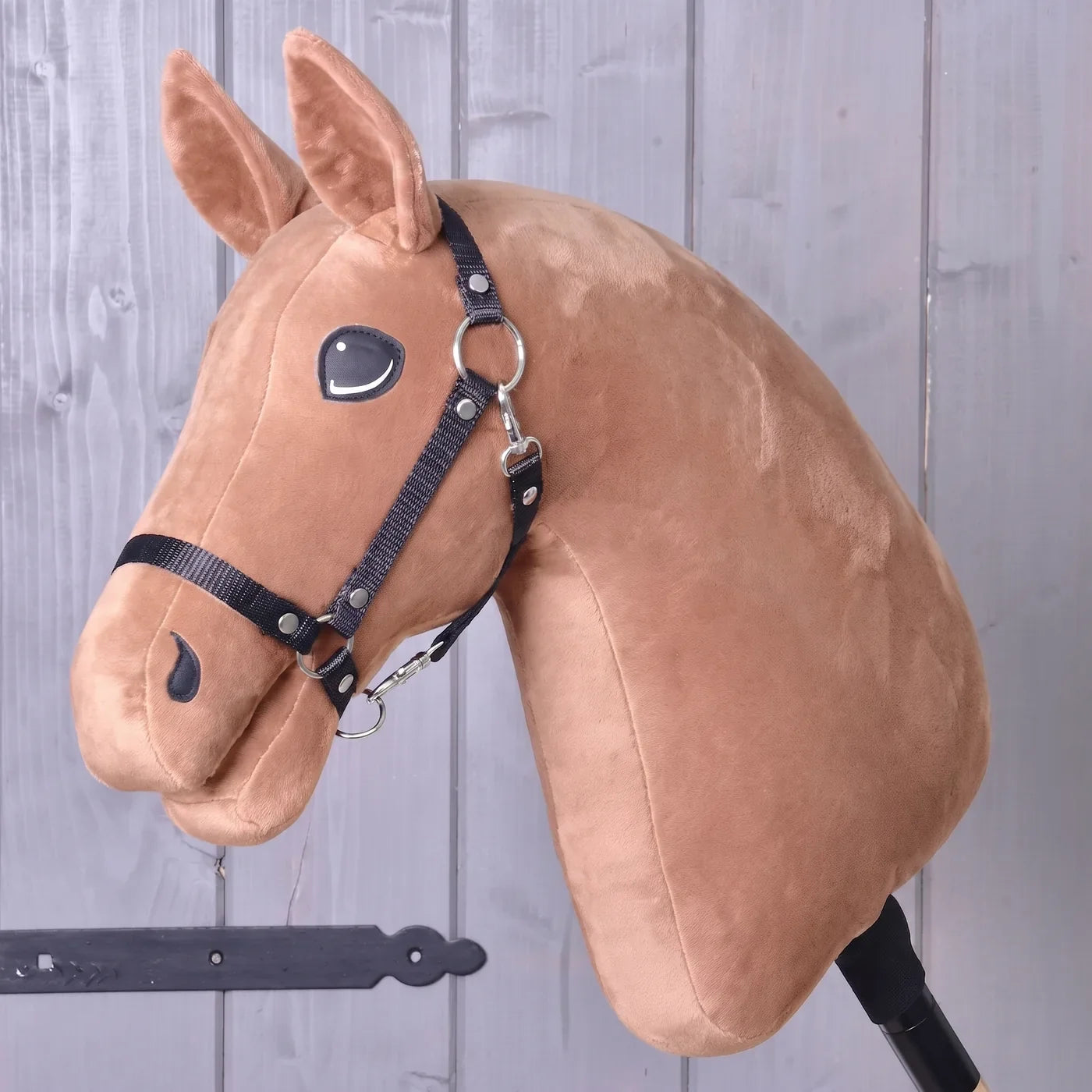 Construis ton cheval de Hobby Horse LarDen — tête de Hobby Horse en peluche avec bride, montée sur un bâton, posée contre un fond en mur de bois, idéale comme inspiration pour créer votre propre cheval unique avec le configurateur « Build your horse » de Hobby Horse LarDen avant de finaliser votre achat.