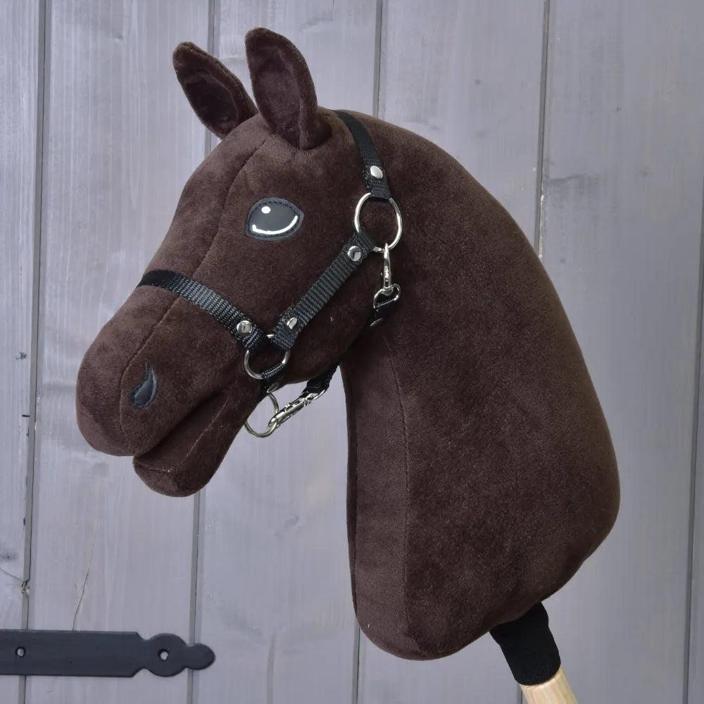 Construis ton cheval, tête personnalisée en peluche pour Hobby Horse, en brun foncé avec bride et rênes noires et crinières blanches remarquables, fixée à un bâton en bois et présentée devant des panneaux en bois gris.