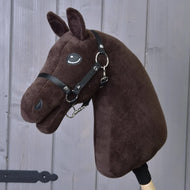 The "Build your horse" plush dark brown hobby horse head, featuring a black harness and bridle and mounted on a wooden stick, is displayed in front of gray wooden boards.