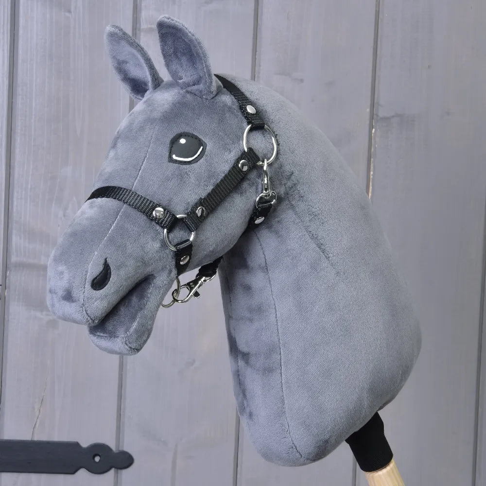 A dark grey “Build your horse” hobby horse by related_to_12441055625556, featuring a black bridle and mounted on a wooden stick, is displayed in front of a wooden wall.