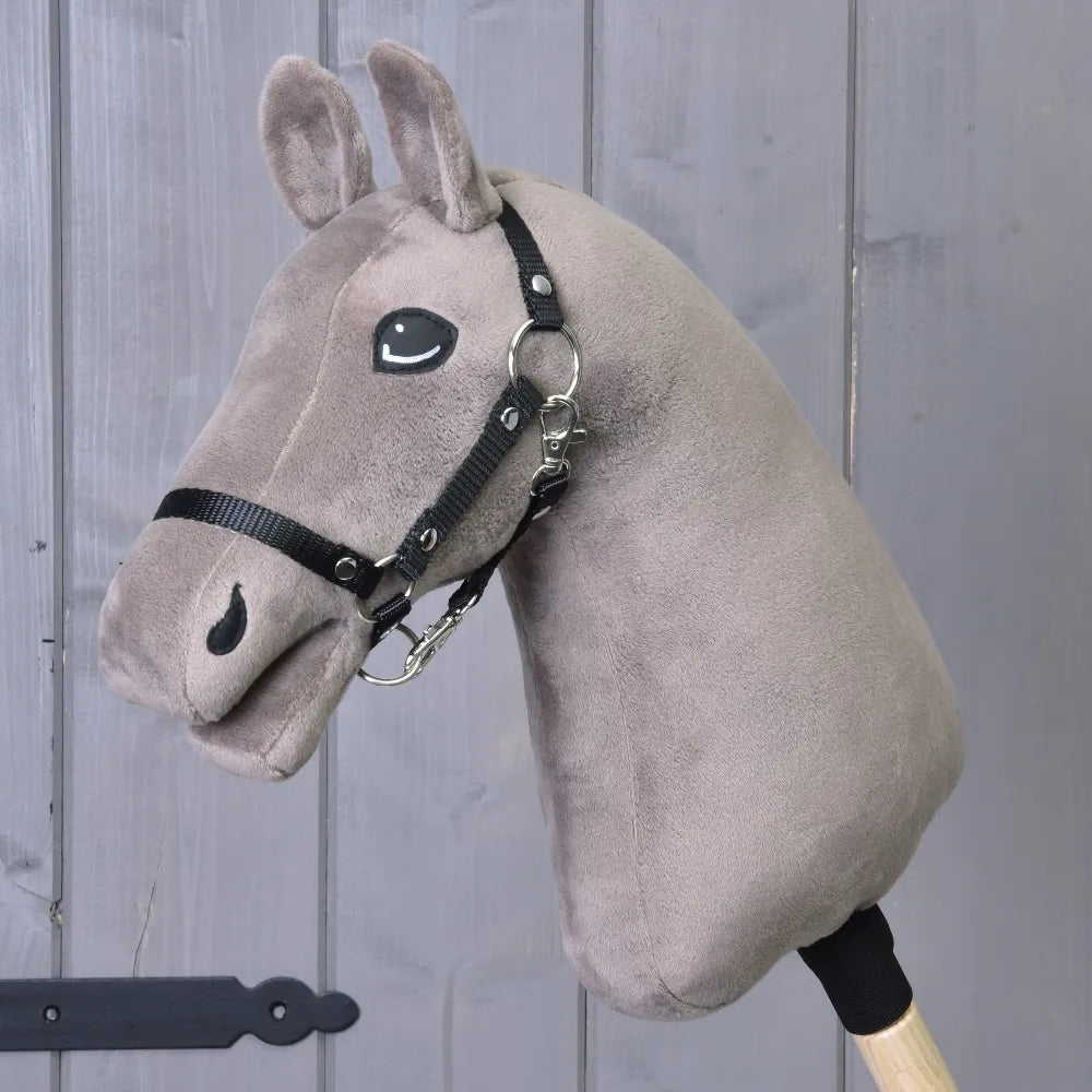 A plush taupe "Build your horse" hobby horse head by related_to_12441055625556, featuring blue eyes and a black bridle, is mounted on a wooden stick and positioned in front of a gray wooden wall.