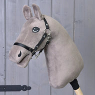 A plush taupe "Build your horse" hobby horse head by related_to_12441055625556, featuring blue eyes and a black bridle, is mounted on a wooden stick and positioned in front of a gray wooden wall.