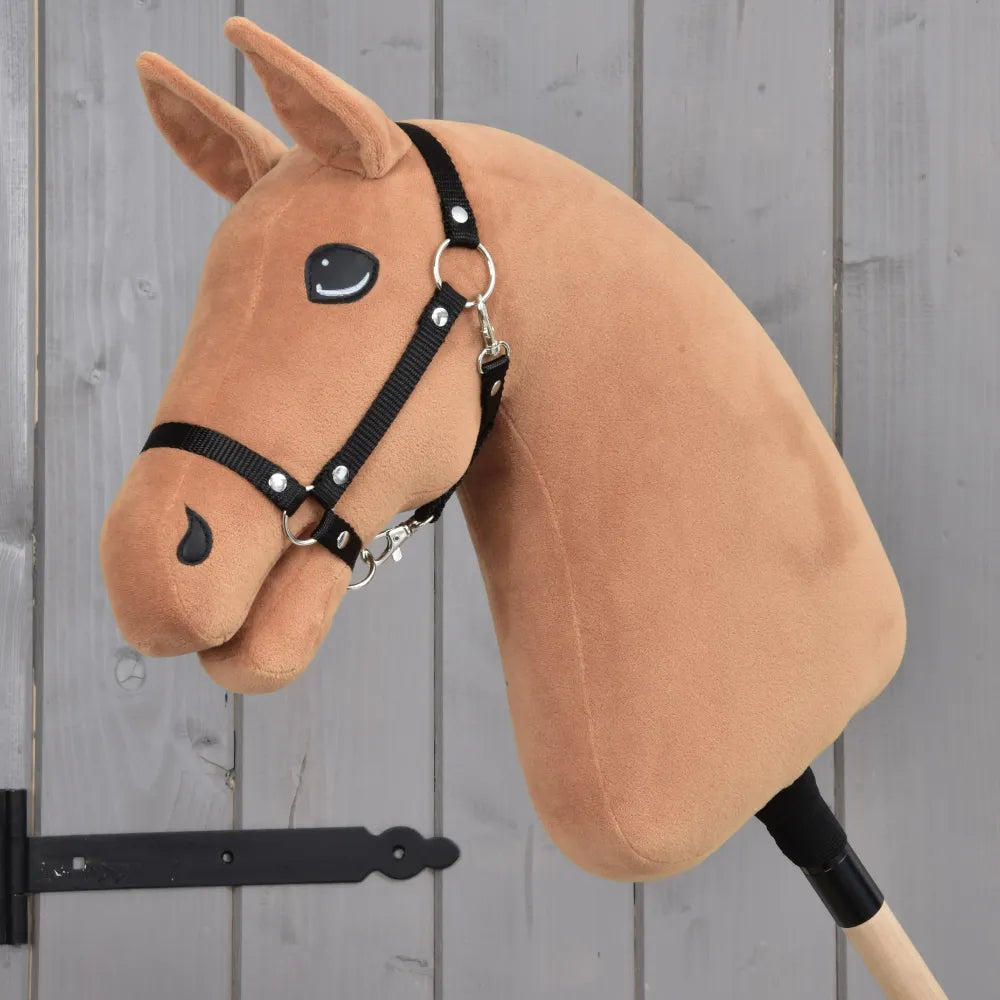 Construis ton cheval, Hobby Horse avec tête personnalisée en peluche brun alezan, bride noire, monté sur un bâton et présenté sur un fond en bois gris avec une charnière métallique noire visible à gauche.