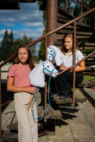 Deux filles avec des Hobby Horses blancs de Hobby Horse LarDen sur un escalier en métal rouillé, le Hobby Horse Lady avec Licol noir devant, tête en peluche blanche et crinière en fil blanc, avec le licol Pastell Blue en similicuir avec fourrure et la longe tricotée Double Blue