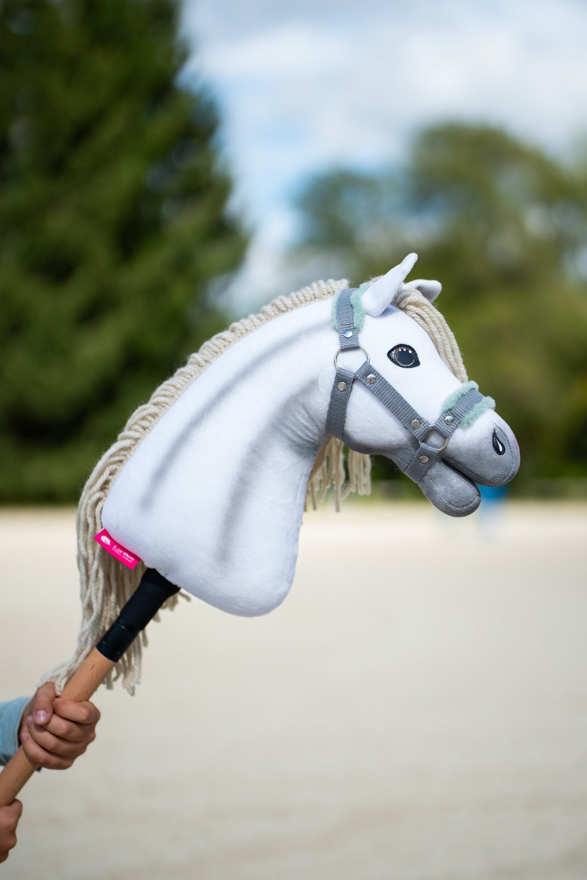 La tête du Hobby Horse Little Lady avec Licol noir de Hobby Horse LarDen est tenue sur une carrière en sable, avec des conifères derrière, peluche blanche, crinière et toupet en fil crème, présenté ici avec le licol gris à fourrure menthe