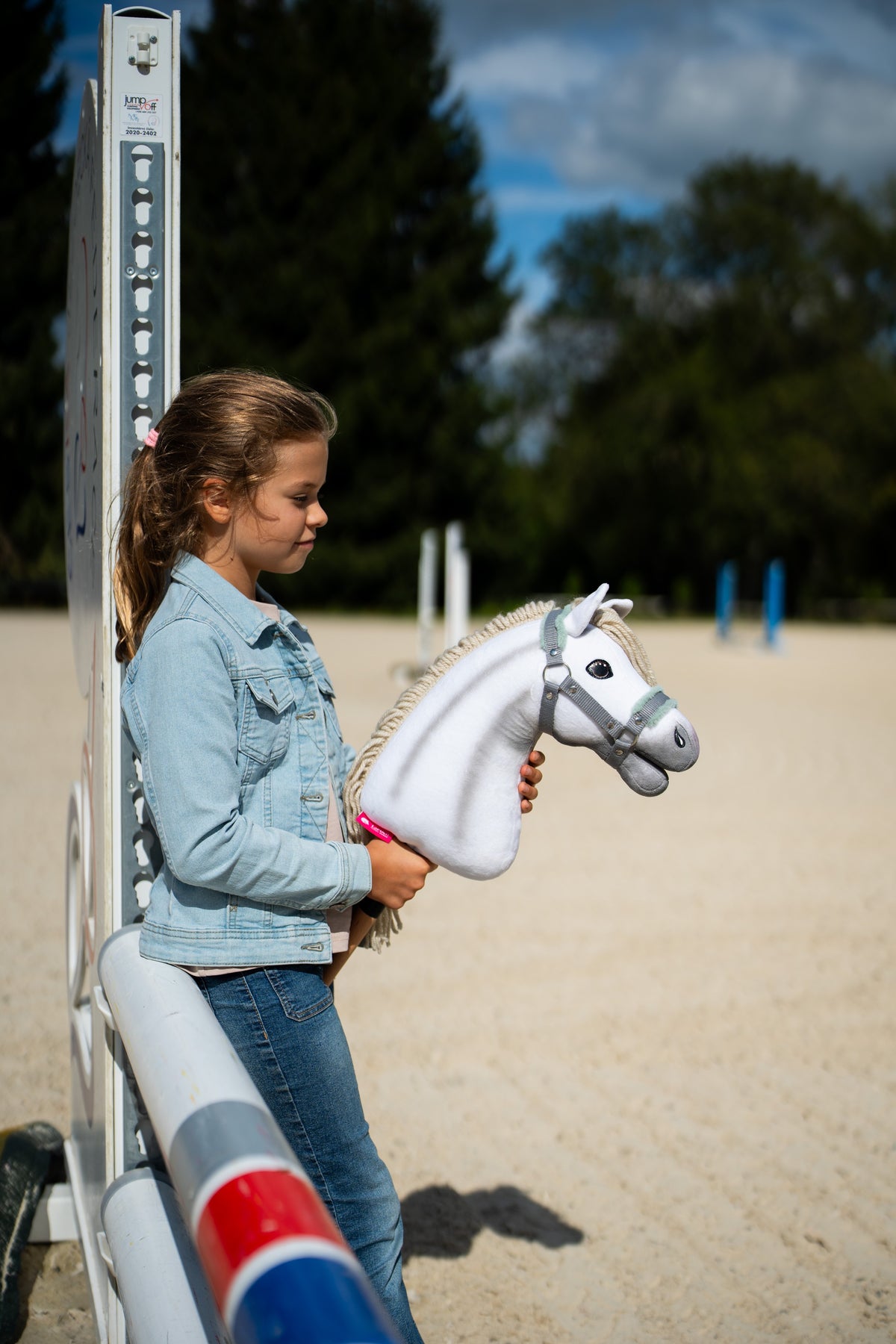 Une fille en veste en jean se tient près d’un chandelier d’obstacle dans une carrière de sable en tenant le Hobby Horse Little Lady avec Licol noir de Hobby Horse LarDen, tête en peluche blanche et crinière en fil crème, avec le licol gris à fourrure menthe
