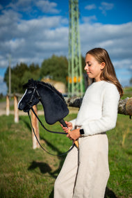 Une fille en pull blanc se tient dans un pré ensoleillé en tenant le Hobby Horse Loki avec Licol noir de Hobby Horse LarDen, tête en peluche noire, liste blanche et crinière en fil noir, présenté ici avec la bride complète Pure Black sans muserolle