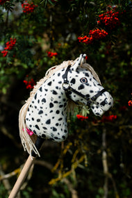 Le Hobby Horse Lord Valentine avec Licol noir de Hobby Horse LarDen devant un sorbier aux grappes de baies rouges, tête en peluche blanche à taches léopard noires, crinière en fil crème et licol noir avec ferrures argentées