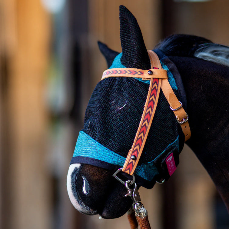 Gros plan sur un cheval portant un masque anti-mouches en maille bleue avec couvre-oreilles et un licol en cuir marron à motif de flèches rouges, rappelant l’équipement de Hobby Horsing. L’arrière-plan est légèrement flou.
