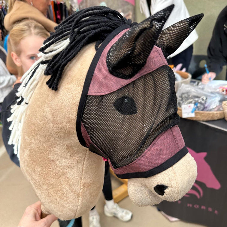 Gros plan sur un Hobby Horse fait main avec masque en maille et crinière en fil noir et blanc. En arrière-plan flou, des personnes et un stand avec des articles sur le thème du cheval illustrent l’esprit créatif du Hobby Horsing.